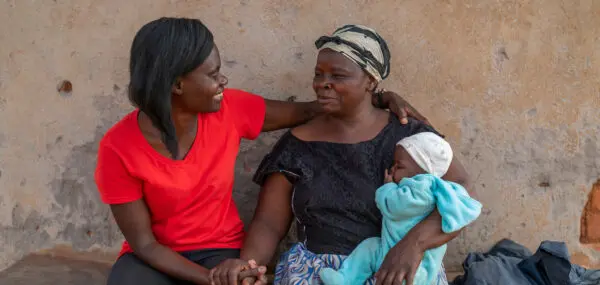 woman in red t-shirt has her arm around a woman with a baby on her arm