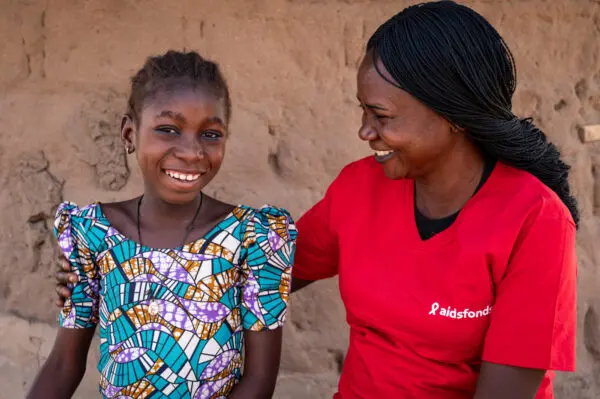 A woman in a red t-shirt has her arm around a young girl in a blue/white dress, smiling, in front of a brown wall