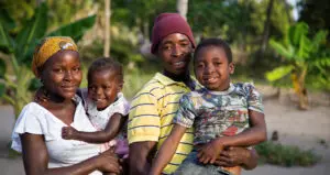 Two adults each carrying a child, the man in a yellow shirt with a dark red hat, the woman with a scarf on her head