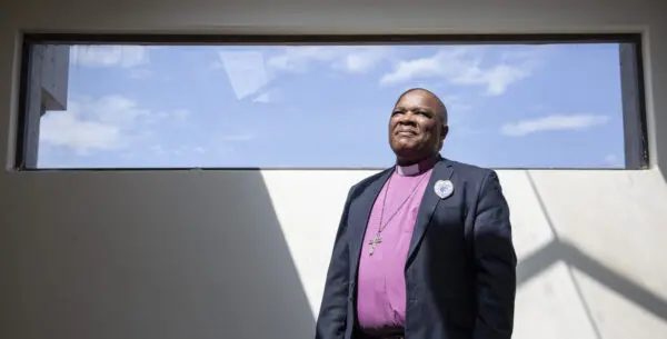 A confident, religious man in a stylish purple suit, standing gracefully in front of a window, exuding elegance and sophistication.