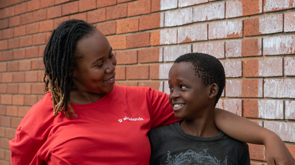 A woman in a red t-shirt has her arm around a boy in a black sihrt: Maselisi and Gift