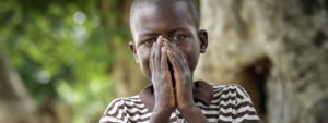 A young boy in a striped shirt stands outdoors with his hands covering his mouth, trees and greenery blurred in the background—a moment of hope inspired by the Paediatric HIV Breakthrough Partnership.