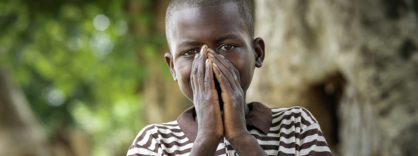 A young boy in a striped shirt stands outdoors with his hands covering his mouth, trees and greenery blurred in the background—a moment of hope inspired by the Paediatric HIV Breakthrough Partnership.
