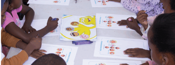 Children sit around a table working on activity sheets with colorful cartoon characters and images, using markers or crayons, while receiving caregiver support for children with HIV in South Africa.