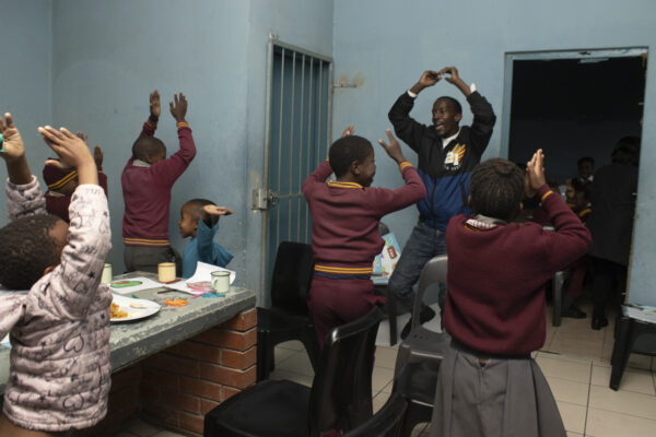 group of children in dark red and grey school uniforms with their arms in the air, following the example of the adult in front of them