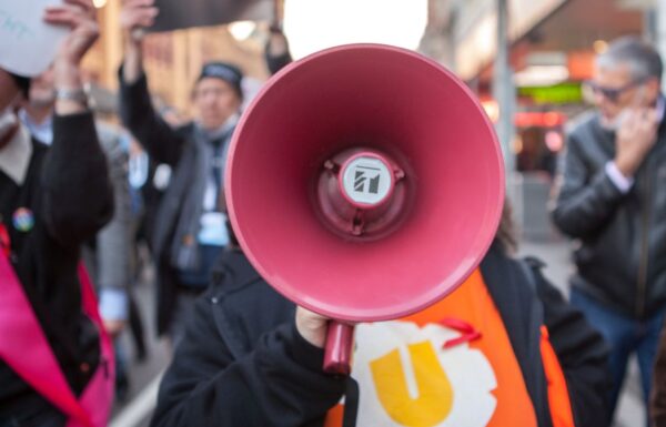 A protester speaking through a megaphone at a demonstration, with others carrying signs in the background.