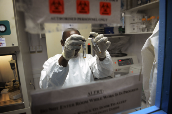 A scientist in protective gear analyzes samples in a high-containment lab, visible through a safety glass window with warning signs.