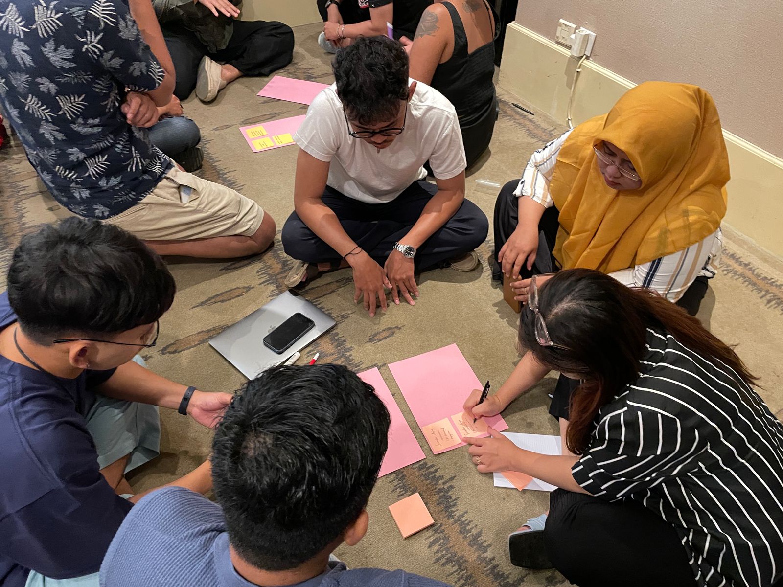 A group of six people is seated on the floor, collaborating on a project with colored paper, markers, and a phone. One person is writing notes while others contribute ideas.