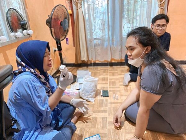 Three people in a room. One person sitting on the floor holding a medical item, another sitting nearby with a mask pulled down, and a third observing. Medical supplies are scattered around.