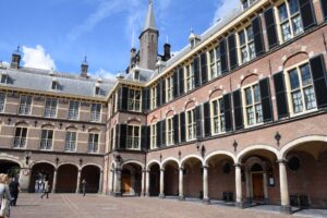 Binnenhof: A historic brick building with arched windows and a clock tower, seen from a courtyard with a few people walking around. The sky is partly cloudy.