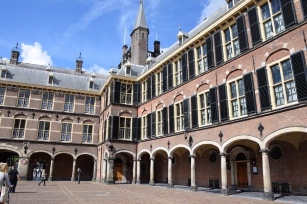 Binnenhof: A historic brick building with arched windows and a clock tower, seen from a courtyard with a few people walking around. The sky is partly cloudy.