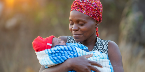 A woman smiles while holding a sleeping baby wrapped in a blue blanket. The woman wears a red headscarf and patterned top, while the baby sports a red hat, all set against a softly blurred background.