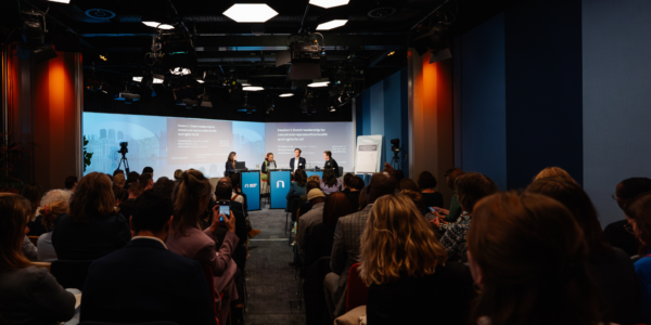 A panel of four experts is seated on stage with a presentation screen behind them, addressing pressing issues in global health to an attentive audience in a dimly lit conference room.