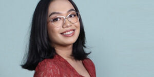 Person with shoulder-length black hair and glasses smiles against a light blue background, embodying the vibrant spirit of an Indonesian LGBTQI+ youth leader, while wearing a striking red outfit.