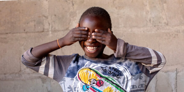 Child smiling and covering their eyes, wearing a colorful skateboard-themed shirt, standing in front of a textured stone wall.