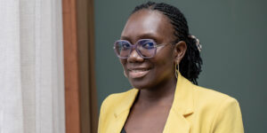 A person wearing glasses and a yellow blazer smiles while standing next to a curtain, against a green background.