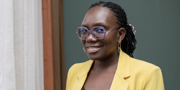 A person wearing glasses and a yellow blazer smiles while standing next to a curtain, against a green background.