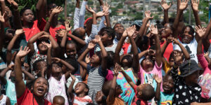 A group of people, including children and adults, raise their hands joyfully outdoors.