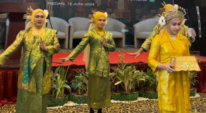 Four people in traditional green and yellow attire perform on a stage with a red carpet and plants. The backdrop displays "Medan, 15 Juni 2024.