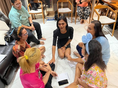 A group of seven people sit on the floor in a circle, engaged in discussion, with papers and pens in hand. Some are seated on chairs, and a sofa is visible in the background.