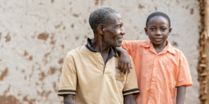 An older man and a young boy stand together. The boy rests his arm on the man's shoulder. They are outdoors, with a textured wall in the background.