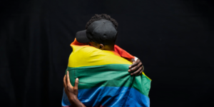 Two people embracing, wrapped in a rainbow flag against a black background, symbolizing unity and hope in the face of adversity, reminiscent of how community support can be as crucial as an emergency fund during times of struggle.