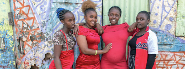 Four young women stand together in front of a colorful, graffiti-covered wall, wearing red and patterned clothing—embodying the spirit of the empowher fund and showing the power of investments needed for a young womenHIV free future.