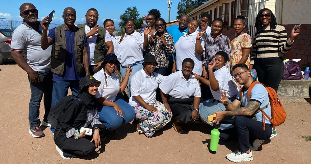 A group of people posing together outside a building on a sunny day. Some wear white shirts; others are in casual attire.