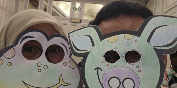 Two people wearing cartoon-style animal masks, one resembling a frog and the other a pig, pose for a photo in an indoor setting.