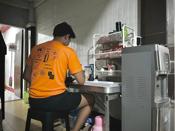 A person in an orange shirt sits on a black stool at a small table, working with a laptop in a kitchen area next to a water dispenser and dish rack, focusing on research about HIV stigma.