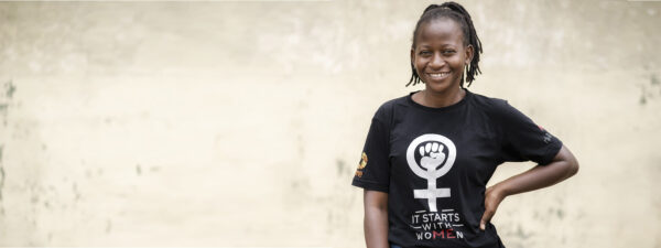 A woman stands smiling against a plain wall, wearing a black t-shirt with a feminist symbol and the words “IT STARTS WITH WOMEN.”.
