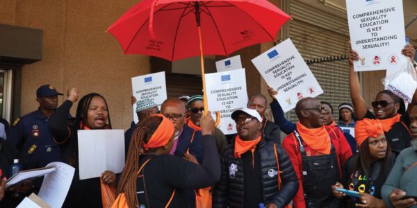 A group of people wearing orange scarves and holding signs advocate for comprehensive sexuality education; one person holds a red umbrella symbolizing sex workers rights and activism.
