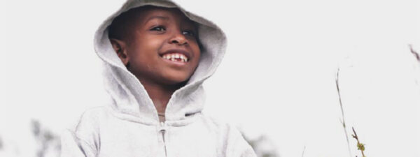 A young child wearing a light gray hoodie smiles while looking off to the side against a bright, blurred background—a hopeful image echoing a sustainable paediatric HIV care in Uganda change story.