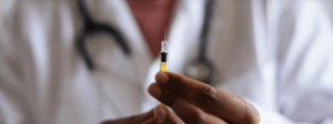 A person wearing a white coat and stethoscope holds a syringe filled with yellow liquid, preparing to administer an injection.