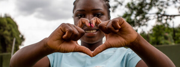 A person wearing a light blue shirt forms a heart shape with their hands, smiling at the camera outdoors with trees in the background.