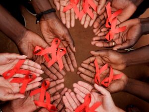 A group of hands arranged in a circle, each holding a red ribbon symbolizing HIV/AIDS awareness.