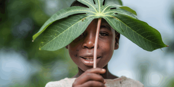 A child holds a large green leaf above their head, using it like a hat, and smiles at the camera.