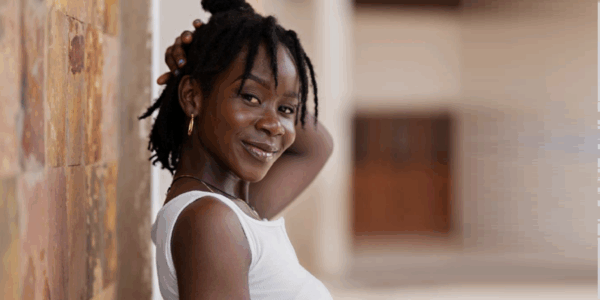 A woman with dark braided hair wearing a white sleeveless top smiles while leaning against a textured wall, with her hand resting on her head.