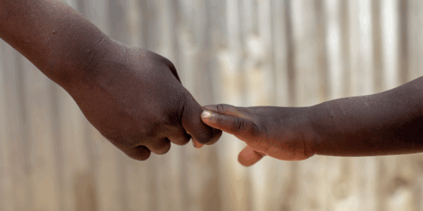 An adult hand holds the finger of a child's hand against a blurred, light-colored background.