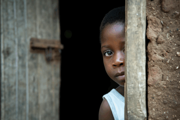 A child in a white tank top stands partially hidden behind a wooden door, looking out with a neutral expression. The background is dark and the wall is made of rough, brown material.