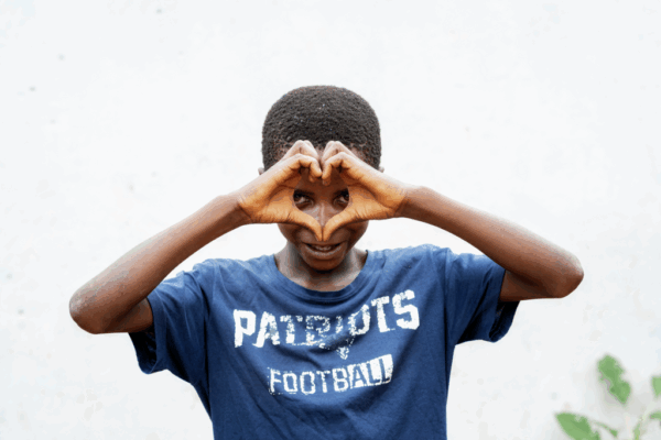 A boy wearing a blue “Patriots Football” t-shirt makes a heart shape with his hands in front of his eyes, symbolizing support for the organisation strategy 2026-2030, standing against a plain white background.