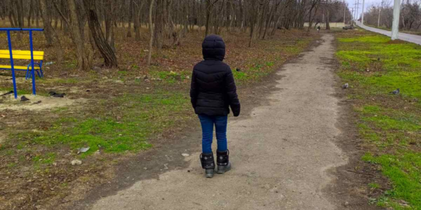 A person wearing a black jacket and rollerblades stands on a dirt path in a park with leafless trees and patches of grass, as if untouched by the war that has reshaped the world beyond these quiet borders.