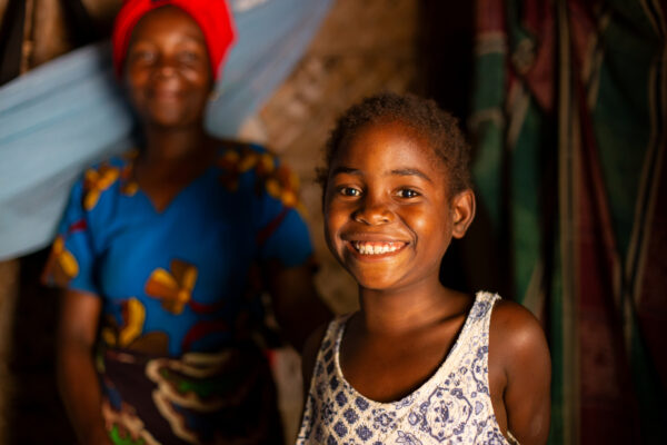 Smiling young girl stands in the foreground with a woman in colorful clothing and headwrap blurred in the background indoors.