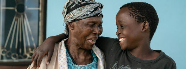 An elderly woman and a young boy smile at each other, with the boy’s arm around the woman’s shoulder, standing in front of a blue wall.