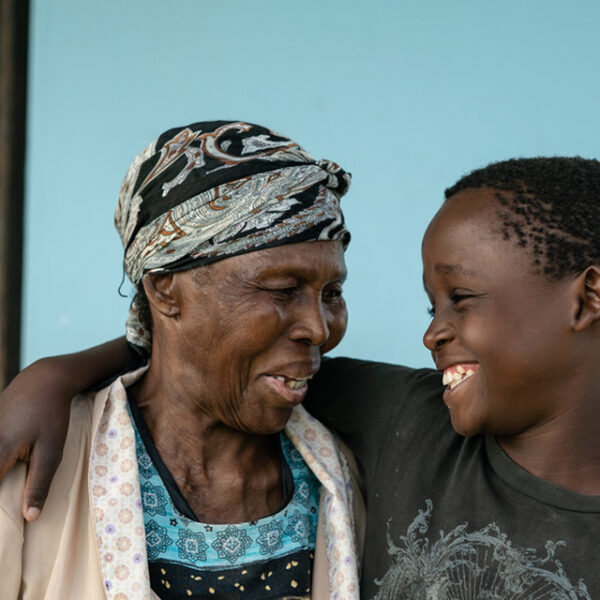 An elderly woman and a young boy stand close together, smiling and embracing in front of a plain blue background.