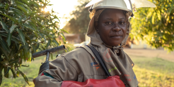 A person wearing protective work clothing and a helmet stands outdoors near trees in daylight, carrying equipment.