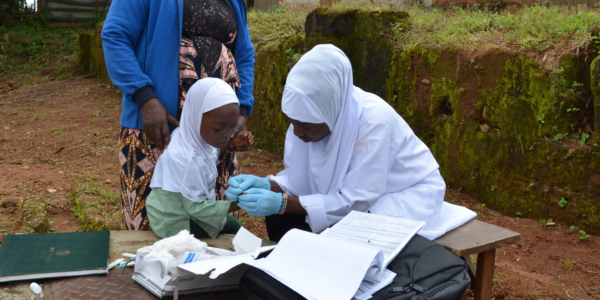 A healthcare worker in white attire administers a medical test to a young girl seated outdoors, while an adult stands nearby. Medical supplies and documents are on the table, reflecting efforts to address paediatric HIV in Nigeria.