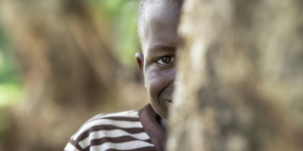 A child wearing a striped shirt stands outdoors, partly hidden behind a tree, with one eye and part of a smile visible.