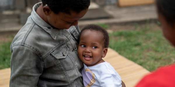 An adult holds a smiling toddler outdoors on a mat. The adult looks down at the toddler, who is looking up and grinning. Another person is partially visible in the foreground.