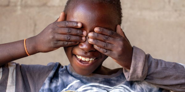 Smiling child covers their eyes with both hands, wearing a grey shirt and an orange bracelet, in front of a plain background.
